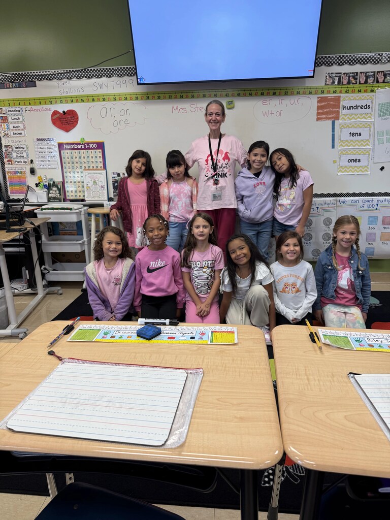 Classroom with teacher, students in pink and white shirts, seated and standing, white boards, desks, and markers.