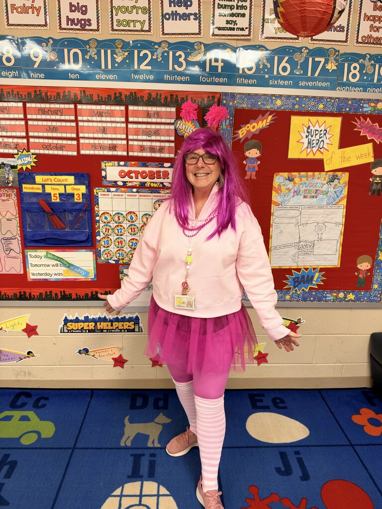Person in pink wig, tutu, and glasses stands in front of bulletin board.