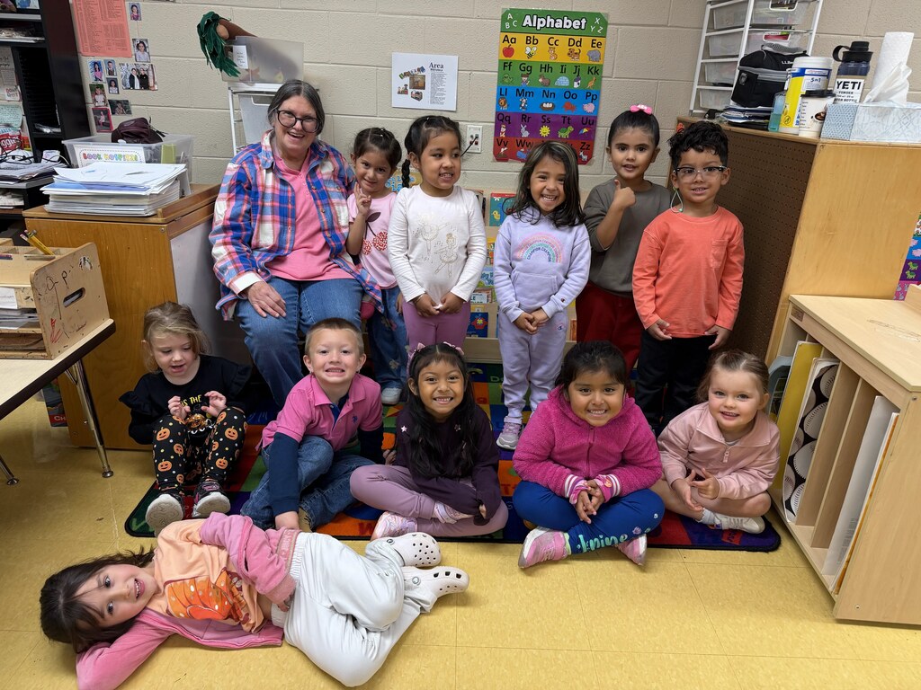 Several young students and a woman sit and stand together on a rug in a classroom.