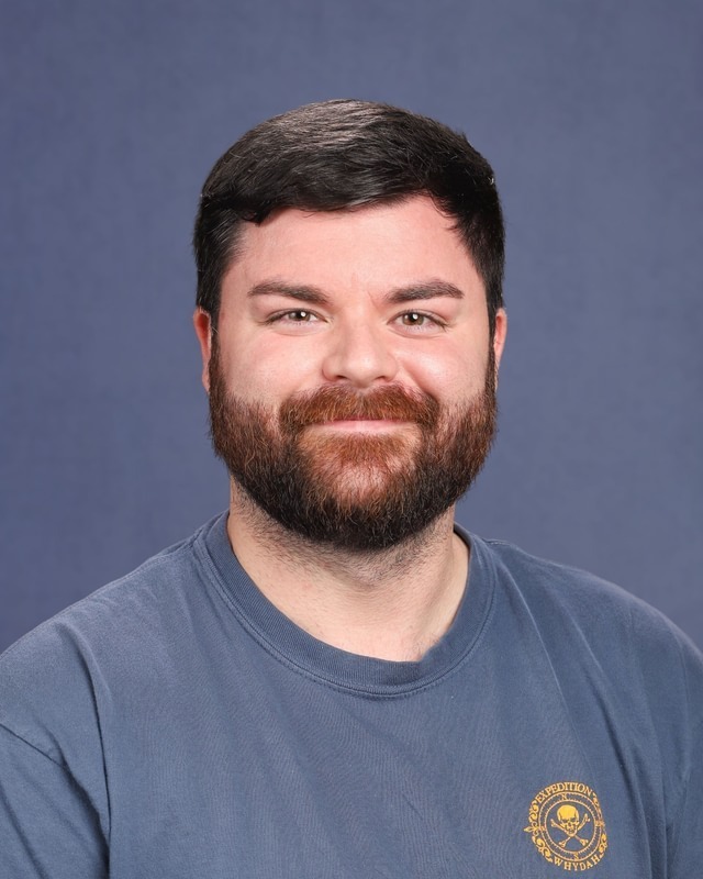 A man with a beard wearing a blue T-shirt with a yellow logo on the left chest area.