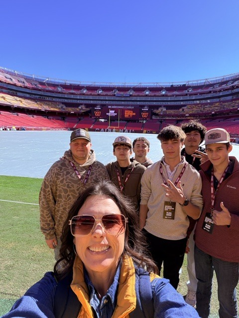 Group of students and adults wearing hats and sunglasses standing on a football field.