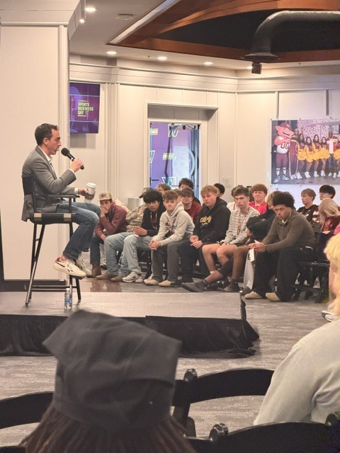 A man on stage with a microphone speaks to a seated audience in a room.