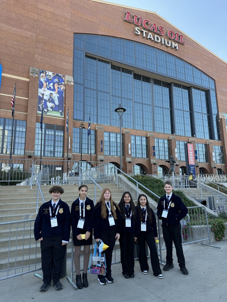 Students in uniforms pose in front of a large stadium with glass walls. A banner hangs above the entrance.