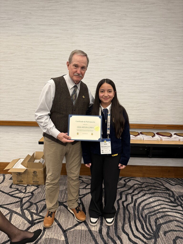 A man and a girl stand holding a certificate. They are in a room with a white wall and patterned carpet.