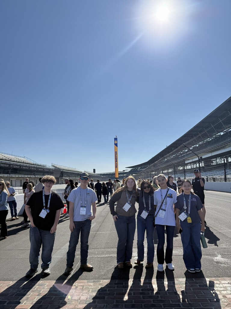Several people stand in a row on a race track with a sunny sky and buildings in the background.