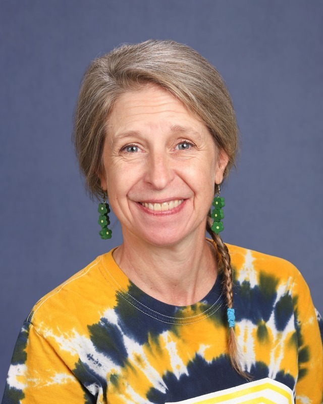 Woman with short gray hair and a smiling face. Wearing a tie-dye shirt and green earrings.