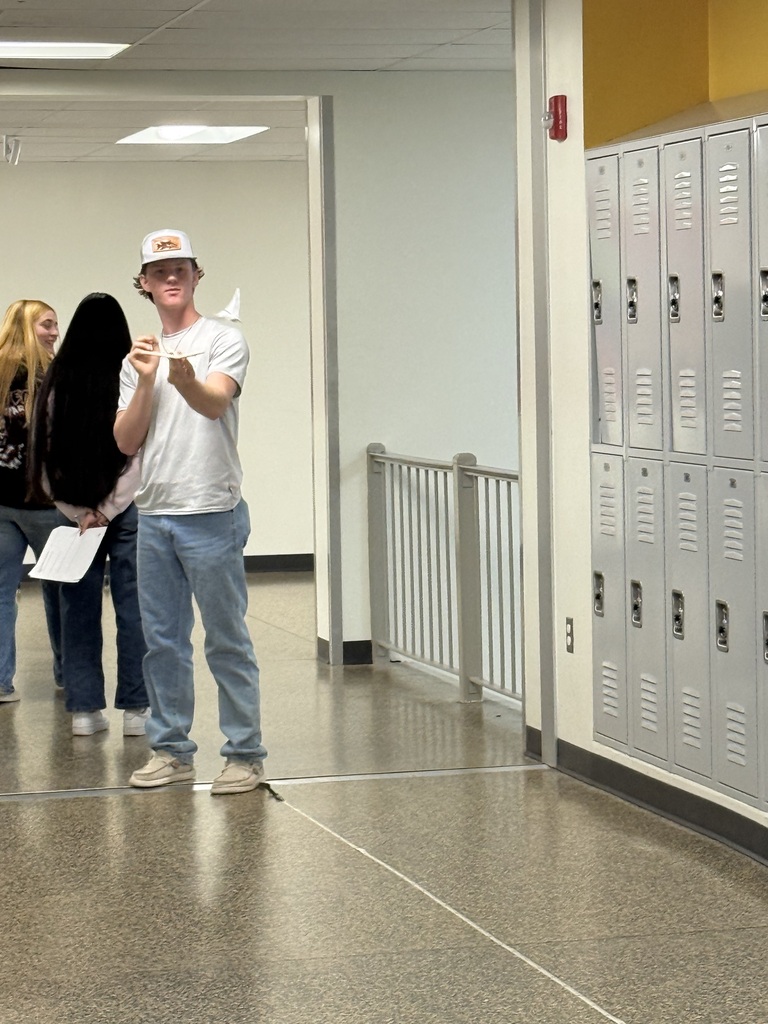 Person wearing hat and sneakers standing in hallway with one hand raised, two people walking behind them, lockers visible on wall.