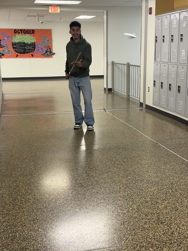 Man with hat and sneakers, holding stick, standing in hallway with lockers, white walls, and floor.