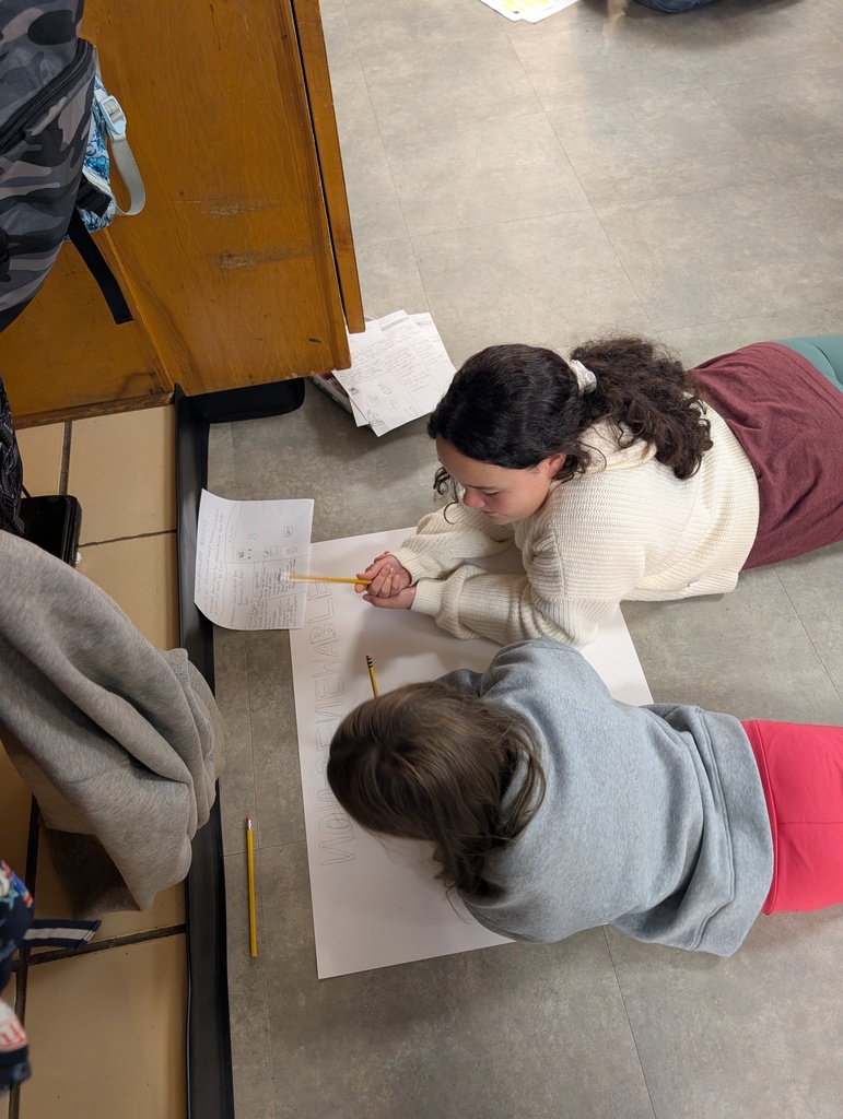 Two students writing on large paper on floor with pencils; one wearing white top, the other in red pants.