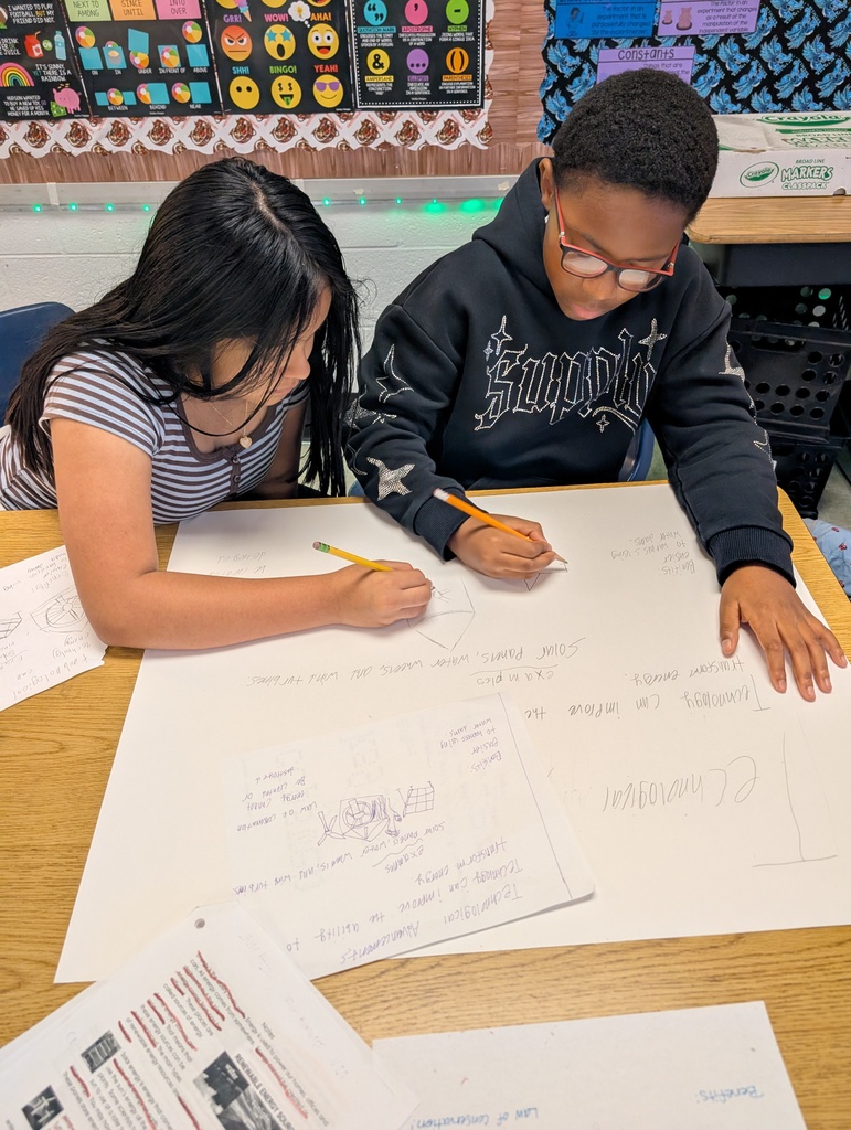 Two students work on a project at a desk with a large piece of paper displaying a diagram.