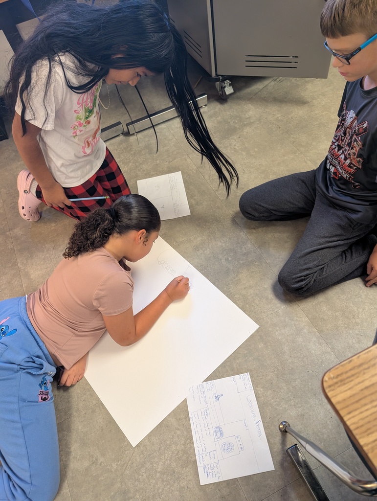 Three students working on a large paper on the floor, with two seated and one standing.