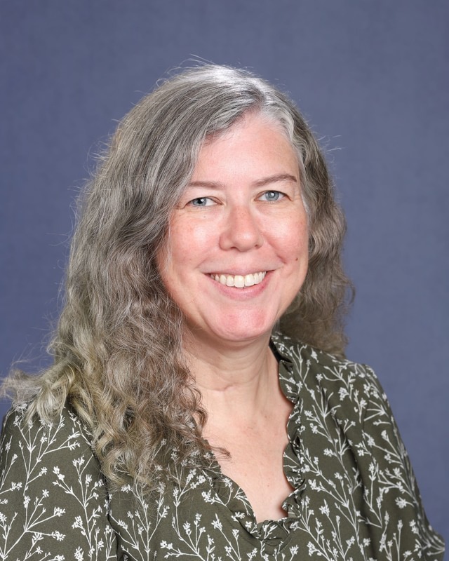 Woman with curly gray hair smiling. Wearing green blouse with white flowers. Blue background.