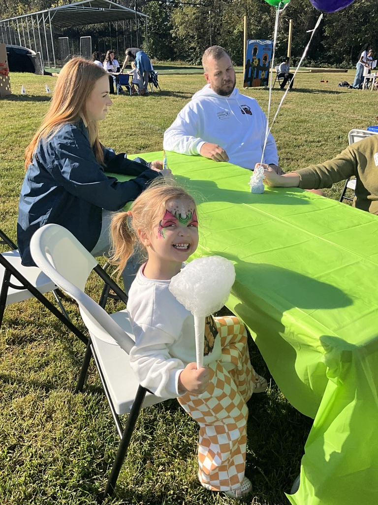 Child holding cotton candy sits in chair at table with adults. Green tablecloth. Trees and building in background.
