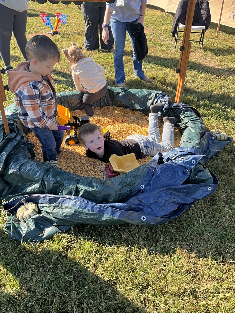 Three children playing in a large container of yellow grains with adults standing in the background.