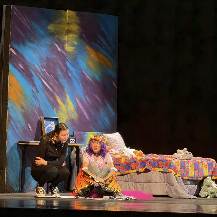 A stage with two performers; one sits on a bed, the other kneels at a desk. Colorful backdrop behind them.