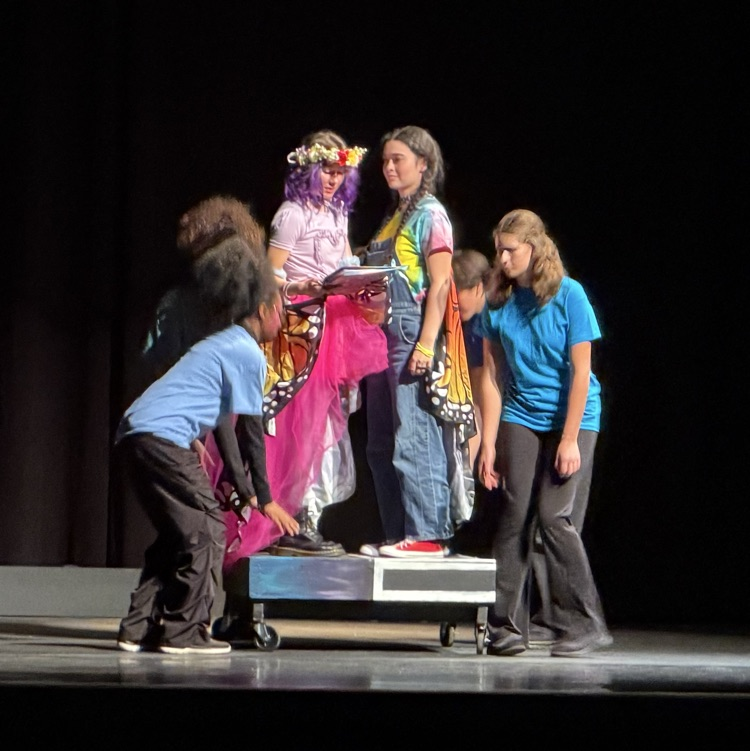 Five children in costumes, one holding a large butterfly prop, are on a stage with a black curtain in the background.