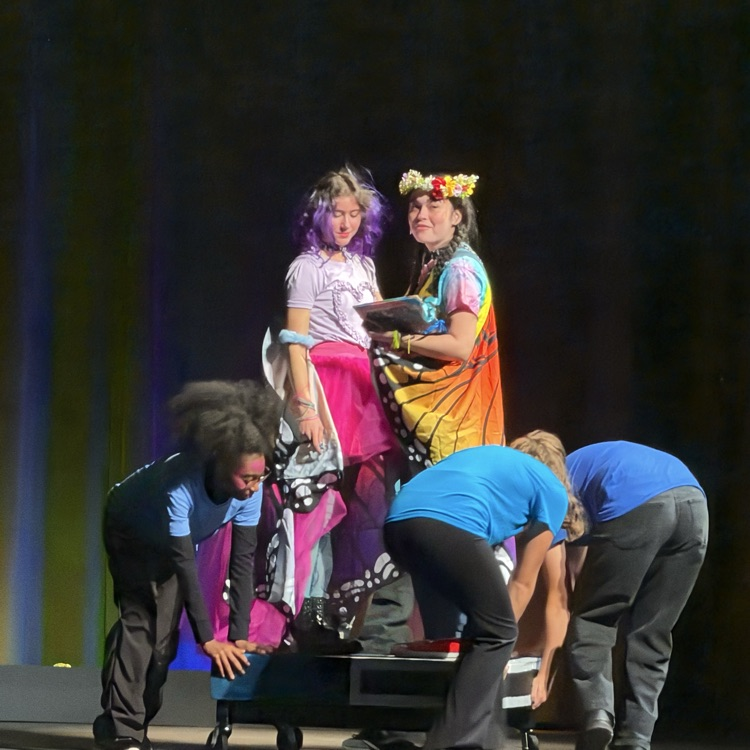 Several students on a stage, two standing in the center, one bent over holding a table, two crouching near table