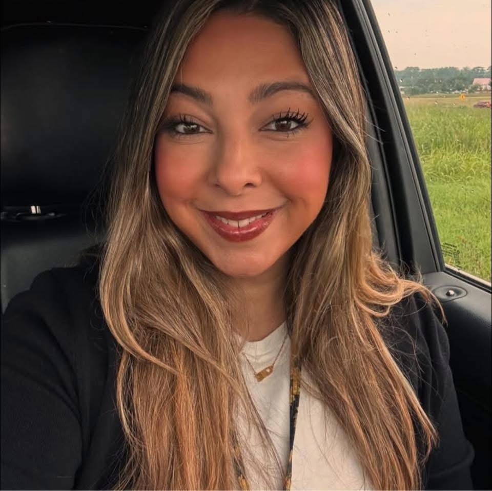 A woman with long hair sits in a car. She smiles and wears a white shirt and black jacket.