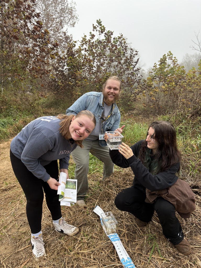 Three students crouch on a grassy hill. Two hold clear containers with water, and one holds a map.