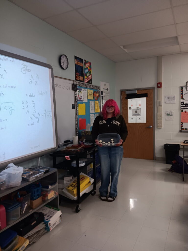 Person with pink hair in classroom, holding container, standing next to whiteboard, bulletin board, and shelves with books.
