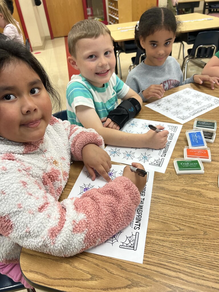 Three children sitting at a table doing a craft with stamps and paper. One child holds a pen.