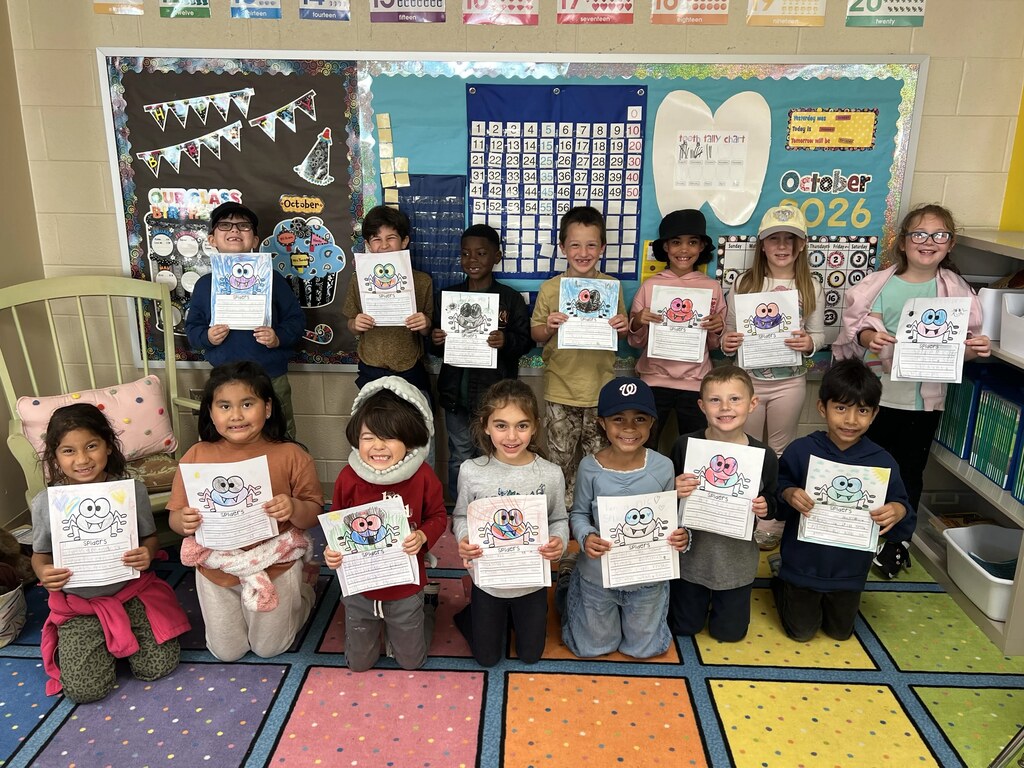 Children in a classroom holding certificates, some wearing hats, colorful floor with bookshelves in background.