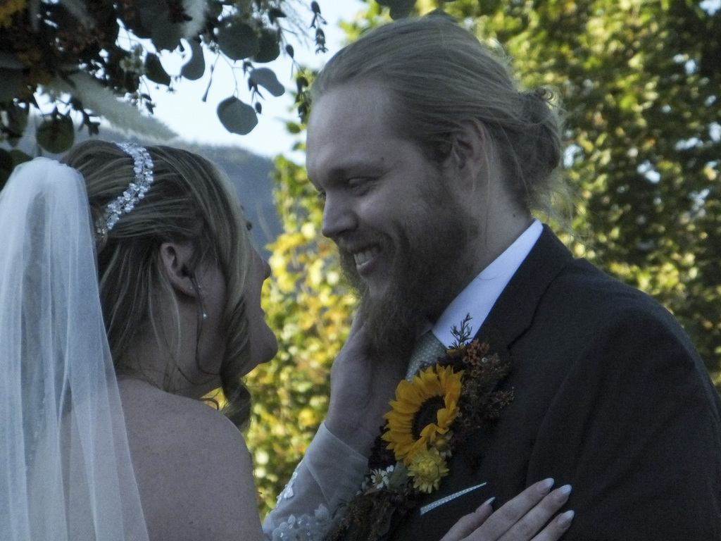 A man in a suit and a woman in a white veil stand close, smiling, outdoors.