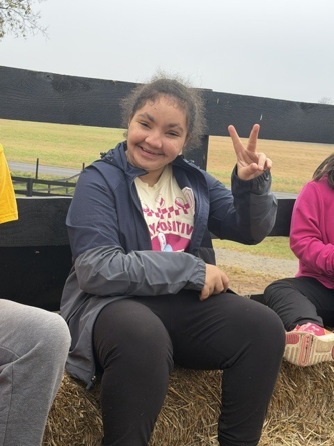 A smiling girl makes a peace sign sitting on hay outside near a fence.