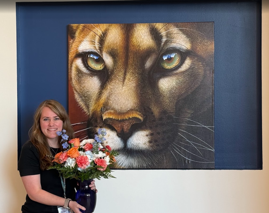 Culpeper Middle School Teacher of the Year Zoe Ingram smiles while holding a colorful bouquet of orange, pink, white, and blue flowers in a dark blue vase. She stands in front of a large, detailed painting of a cougar's face featuring striking green eyes, displayed against a navy blue wall.