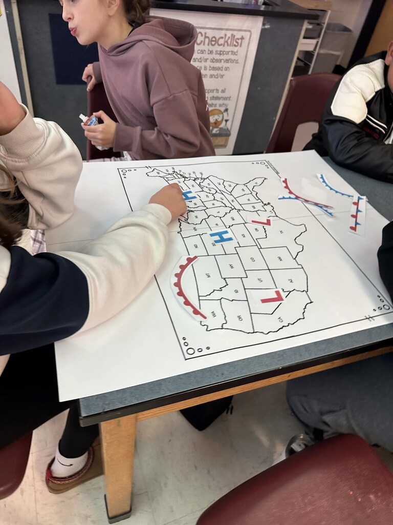 Students seated at a table mark a map with colored markers; one holds a marker.