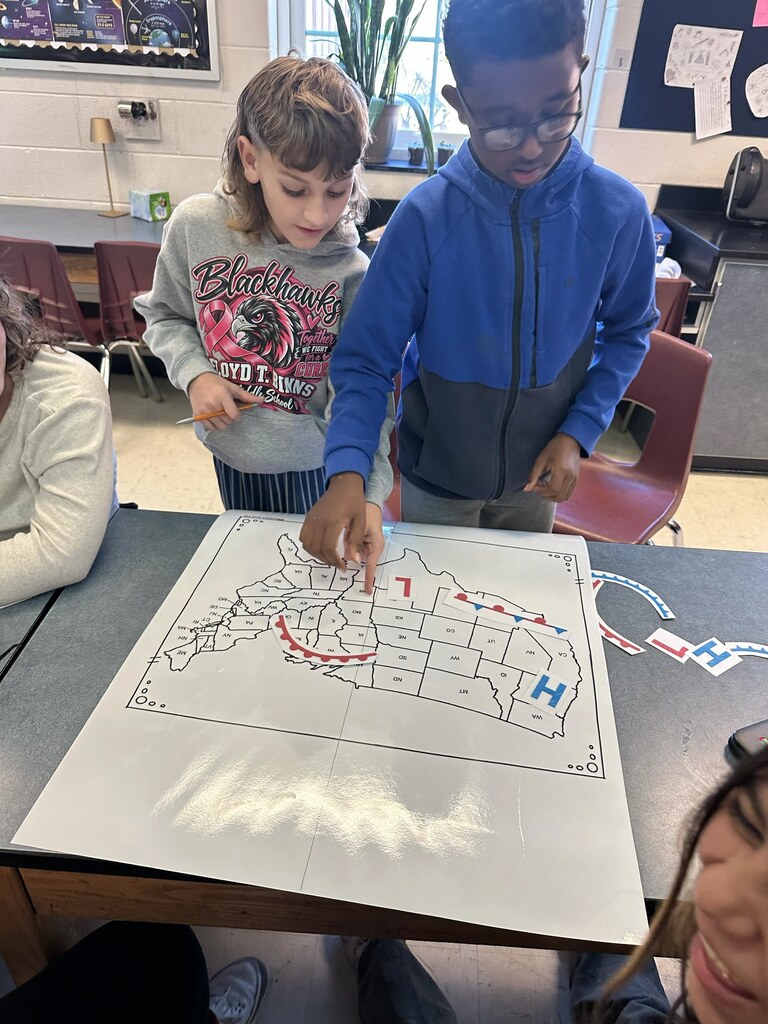 Two kids are standing by a table, pointing to a map with a white outline.