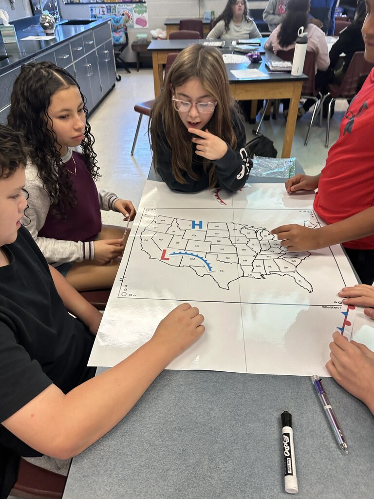 Students at a table, collaborating on a large map of the United States. One student points at the map.