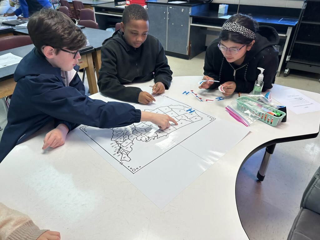 Three students seated at a table, one pointing to a map and others writing on it.