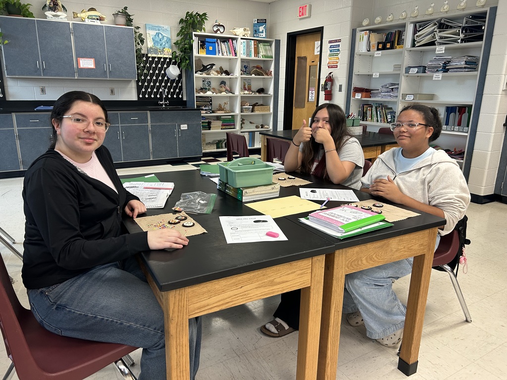 Three students sitting around a table with papers and objects in a classroom with shelves.