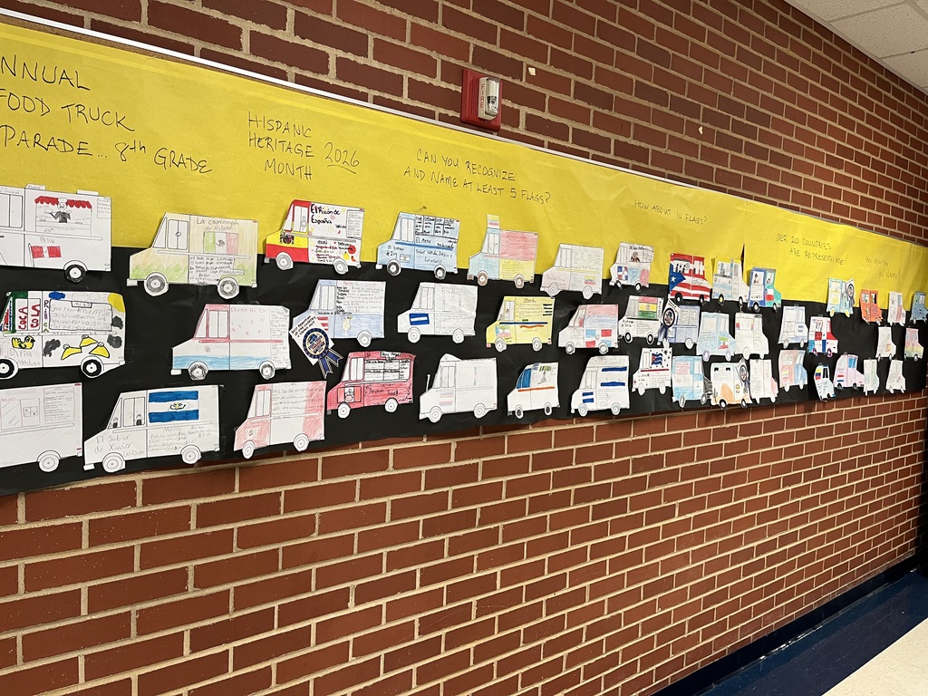 A yellow banner with text is attached to a brick wall. Banner contains drawings of vehicles in a parade.