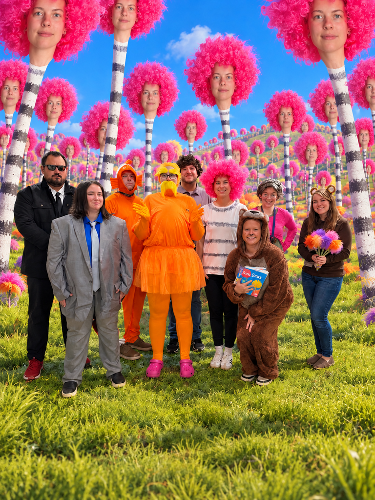 A group of nine CMS staff members and students dressed as characters from Dr. Seuss’s The Lorax pose together in a colorful Truffula Tree forest. Costumes include the Lorax, the Once-ler, a fish, a bear, and other whimsical characters. The background features bright pink, fluffy treetops with faces, striped tree trunks, and a vibrant, grassy landscape.