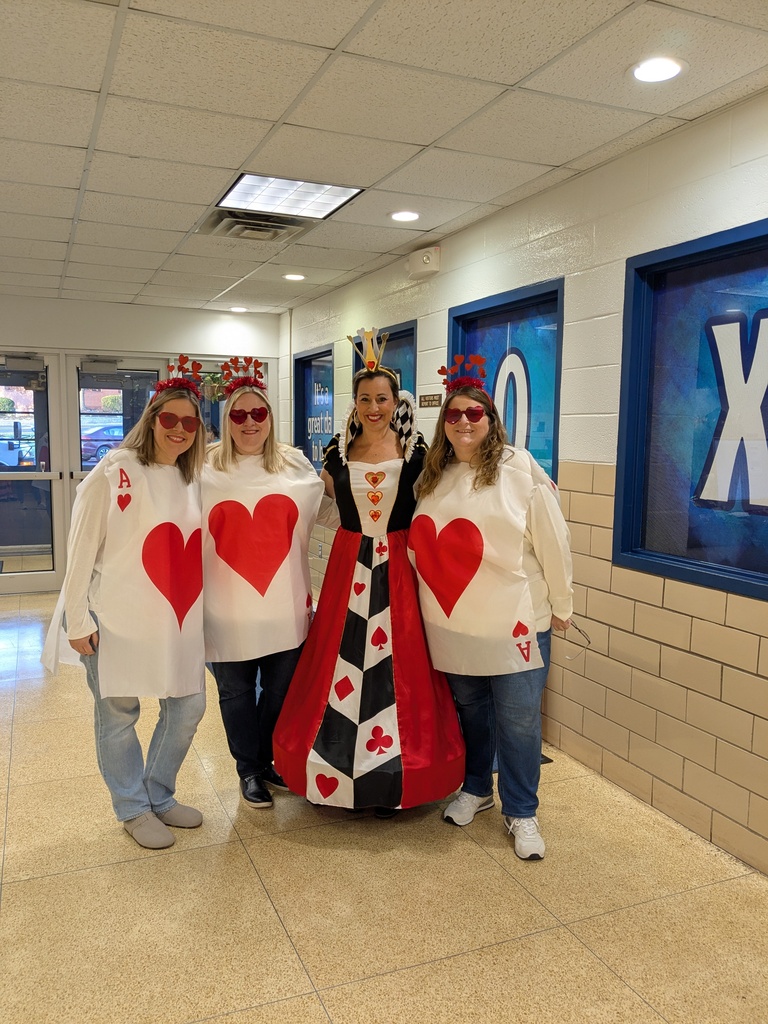 Four people dressed in playing card costumes stand together in a school hallway. The walls have blue signs with white letters.