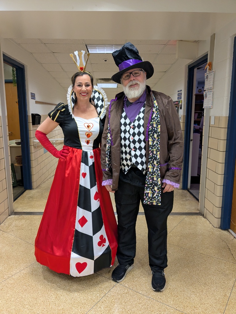 A man and woman stand in a tiled hallway, dressed in costumes. The woman wears a costume with a red and black color scheme. The man wears a purple jacket and hat.