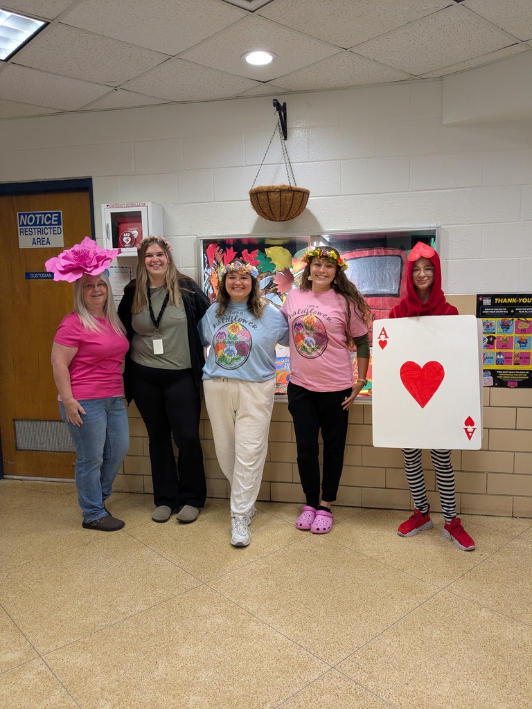 Five women pose in a room. Each wears a pink top. One wears a hat. Three wear accessories to resemble cards.