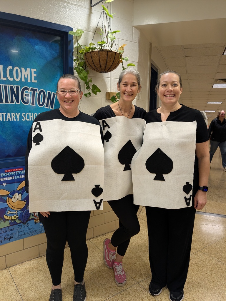 Three women in black hold up signs with spades; one wears pink shoes. Behind them, a wall has blue signage with text.