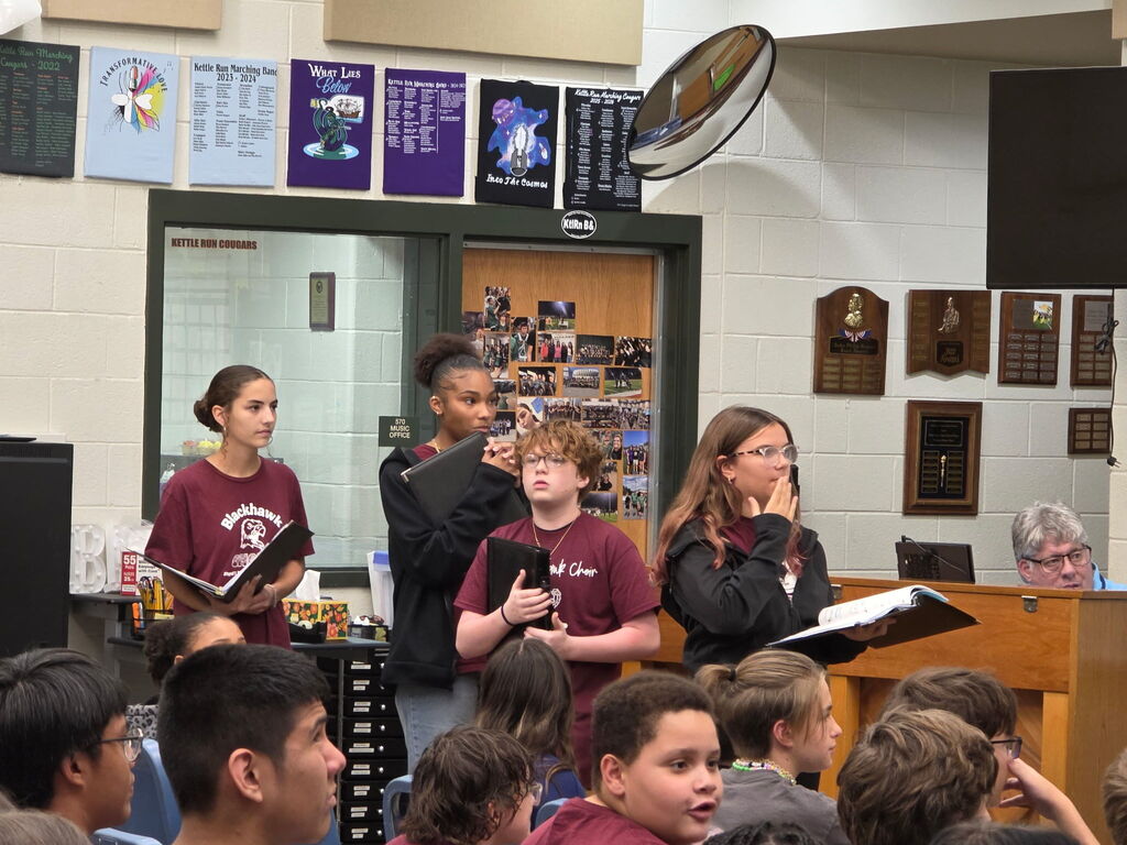 A group of students in maroon shirts stand in front of a crowd, holding books.