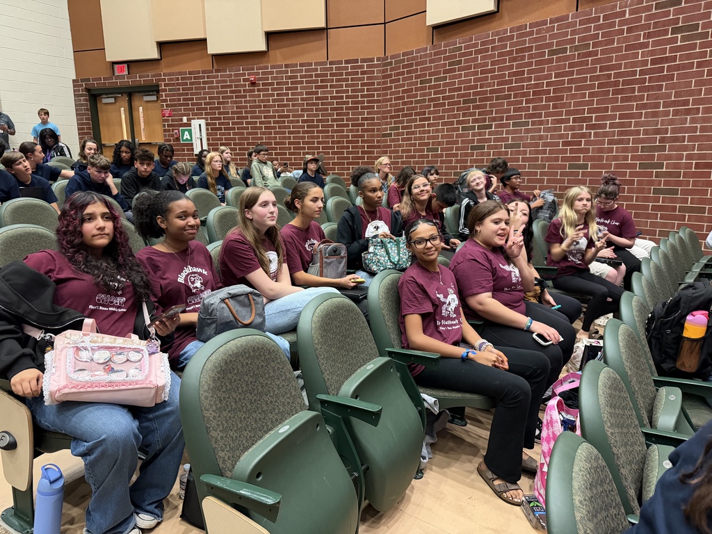 Students in maroon shirts sitting in chairs in a classroom with brick walls.