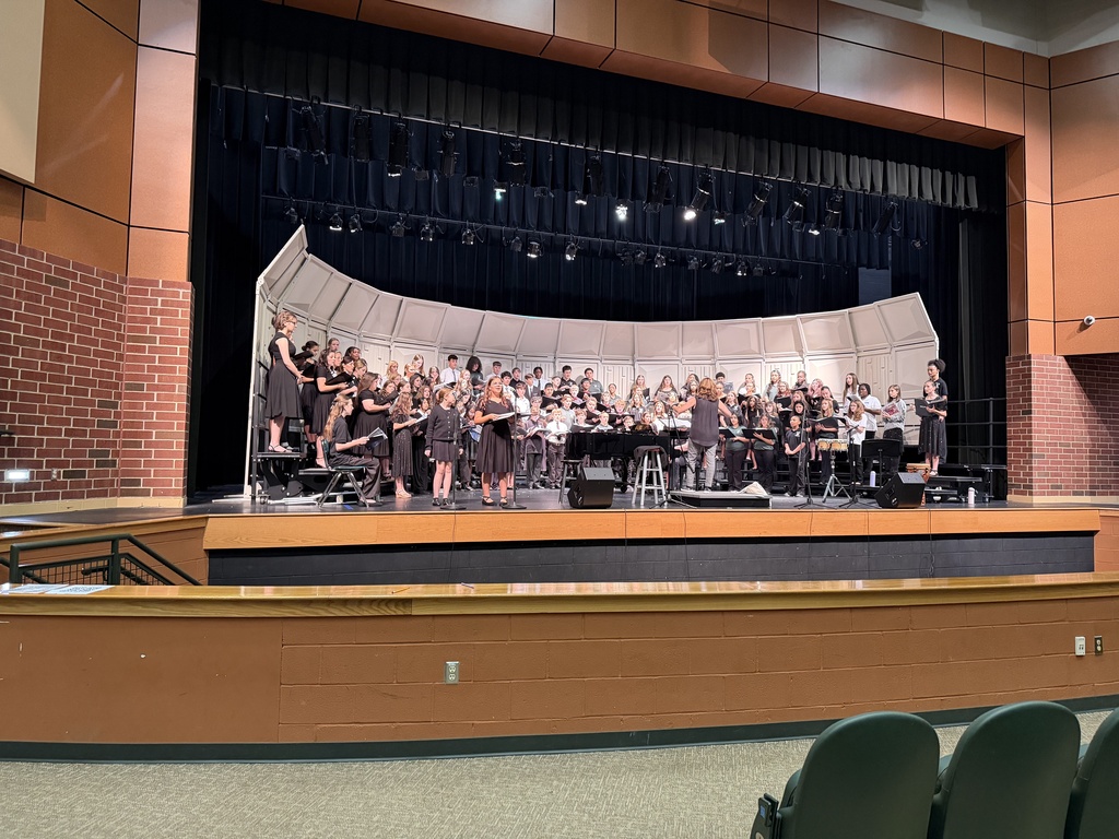 Choir on stage with a white curved backdrop; audience chairs in front; empty auditorium with brick walls.