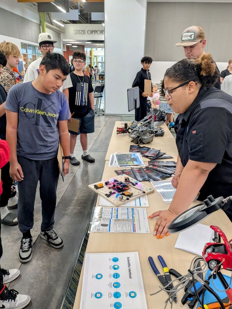 Students examine a table of tools and materials while a woman speaks with them.