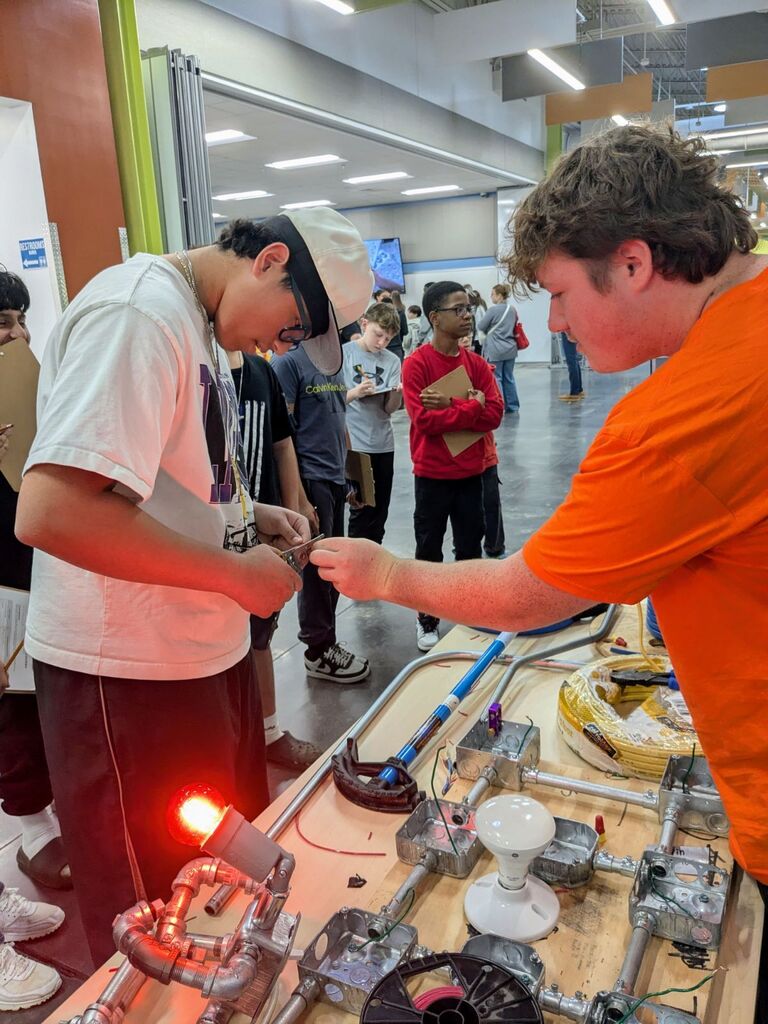 Two students work on an interactive electrical project with wires, lights, and other equipment.
