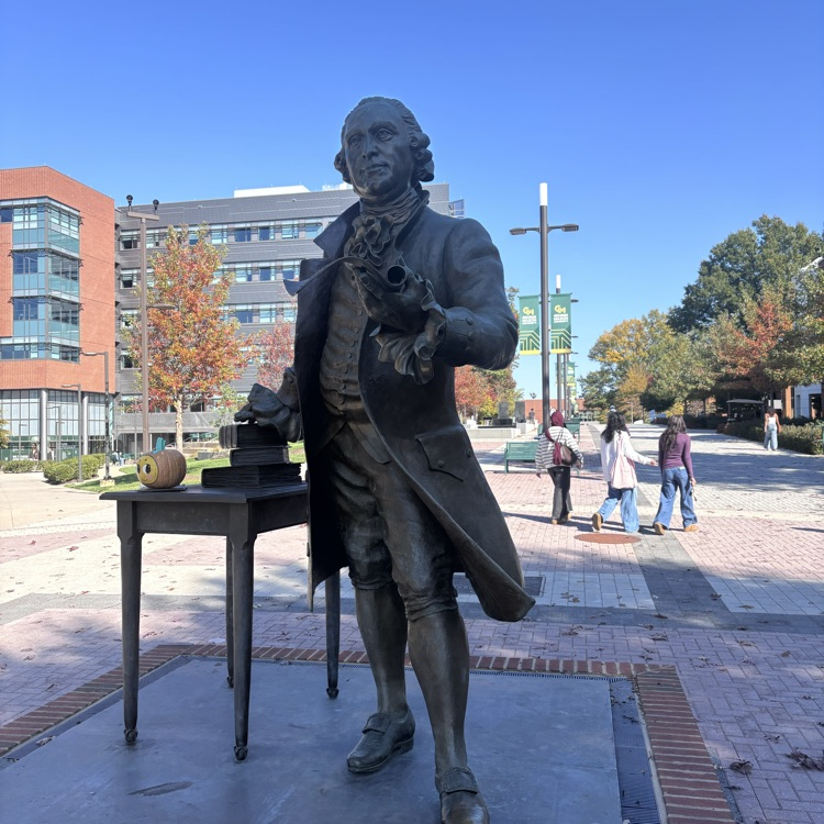 Statue of man wearing a coat stands on platform with table. People walk in background.