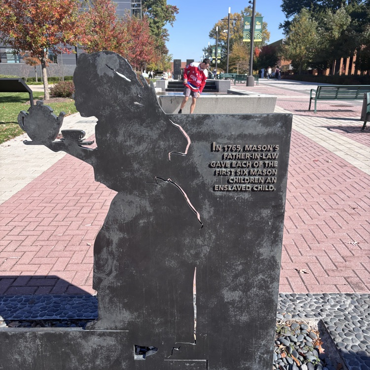 A memorial with an angel holding a cup stands on a brick walkway. A person walks on the path behind it.