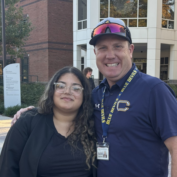 Two people stand smiling in front of a building with a tree, a person, and a sign.
