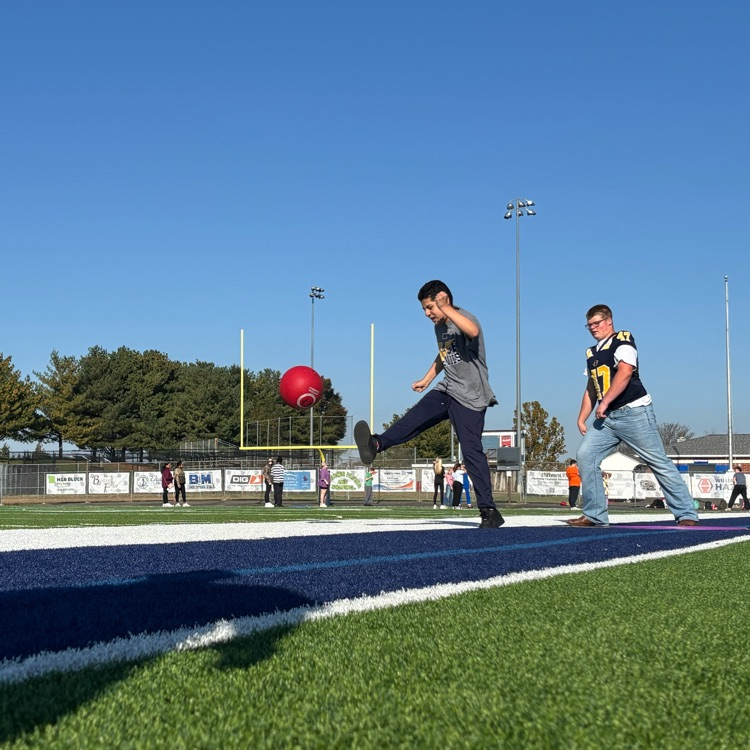 Two people kicking a red ball on a blue and green field. Trees, street lights, and a fence in background.