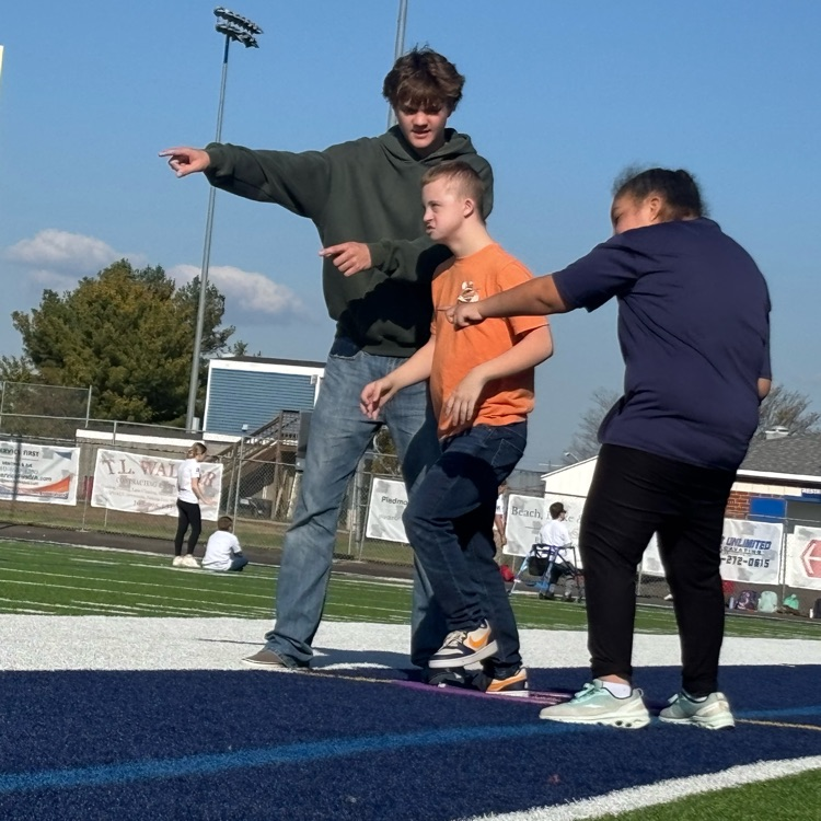A group of people stand on a blue track with white lines. An adult points to a boy.